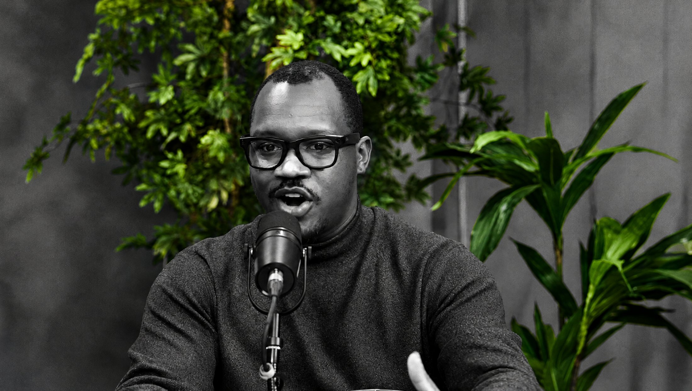 Raheem Manning laughing at the microphone, in black and white with the plants behind him in green