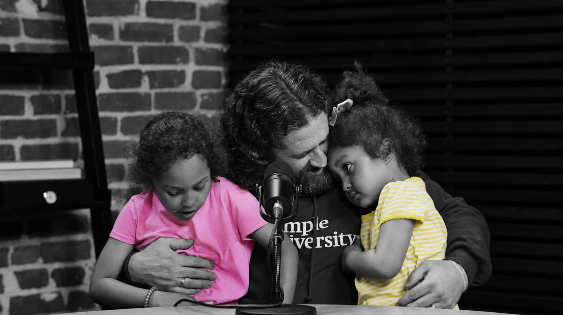 Nathan Luce with his daughters Lulu and Faith on his lap at the microphone, in black and white with the girls' pink and yellow shirts in colour