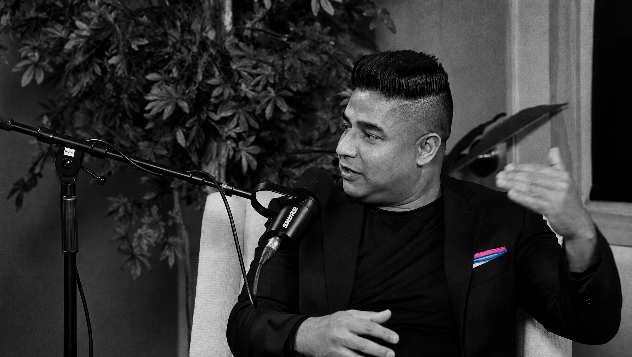 Ajay Raju leaning in mid-gesture, in black and white with his pink and blue pocket square in colour