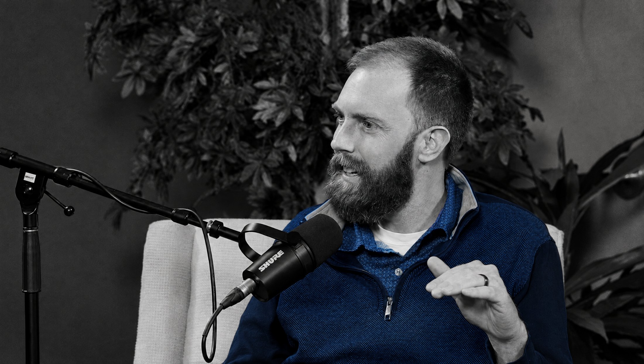 A host mid-sentence at the microphone, hand raised, in black and white with his blue quarter-zip sweater in colour