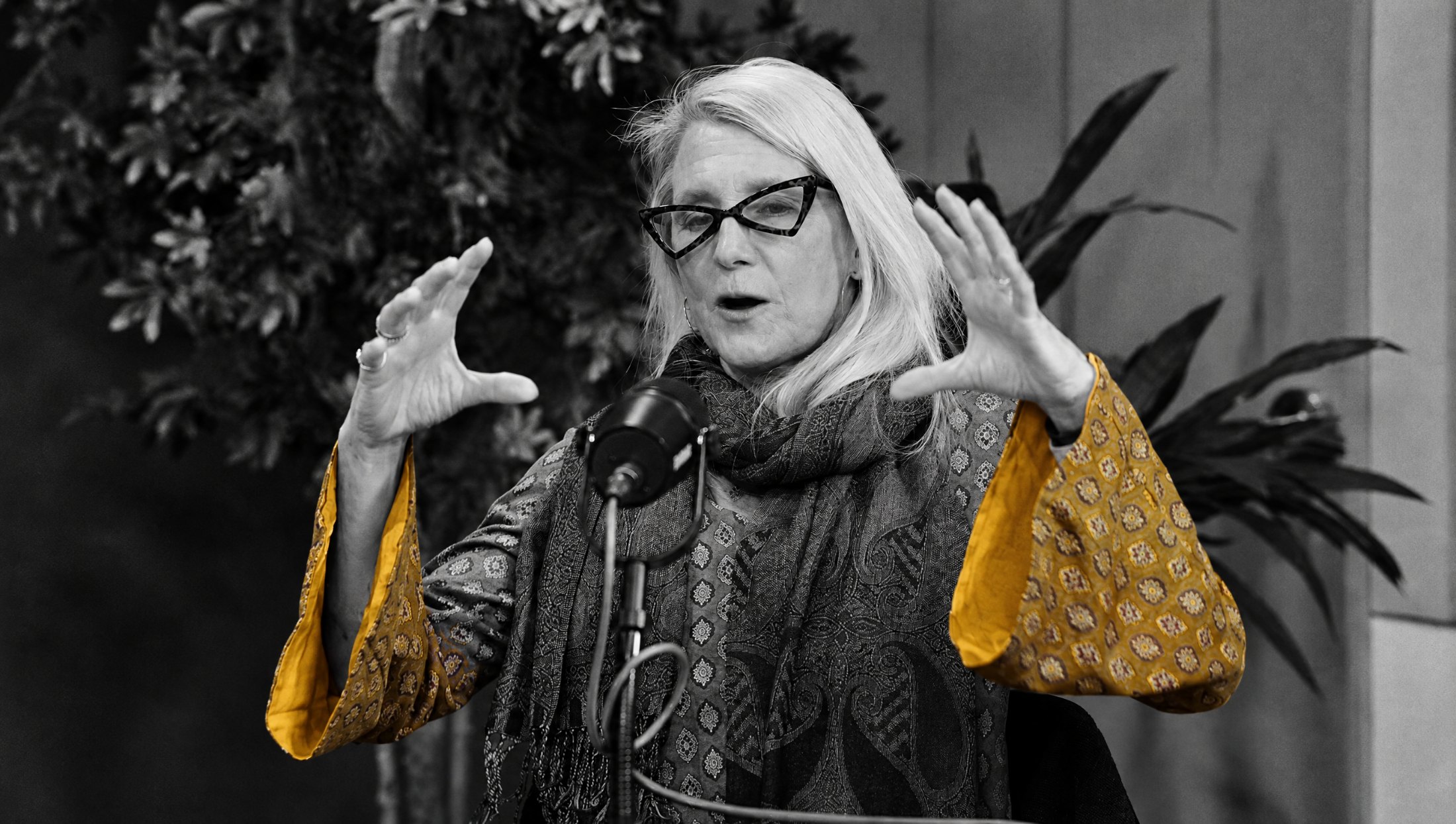 Dr. Lynne Andersson with both hands raised mid-explanation, in black and white with the gold sleeves of her jacket in colour